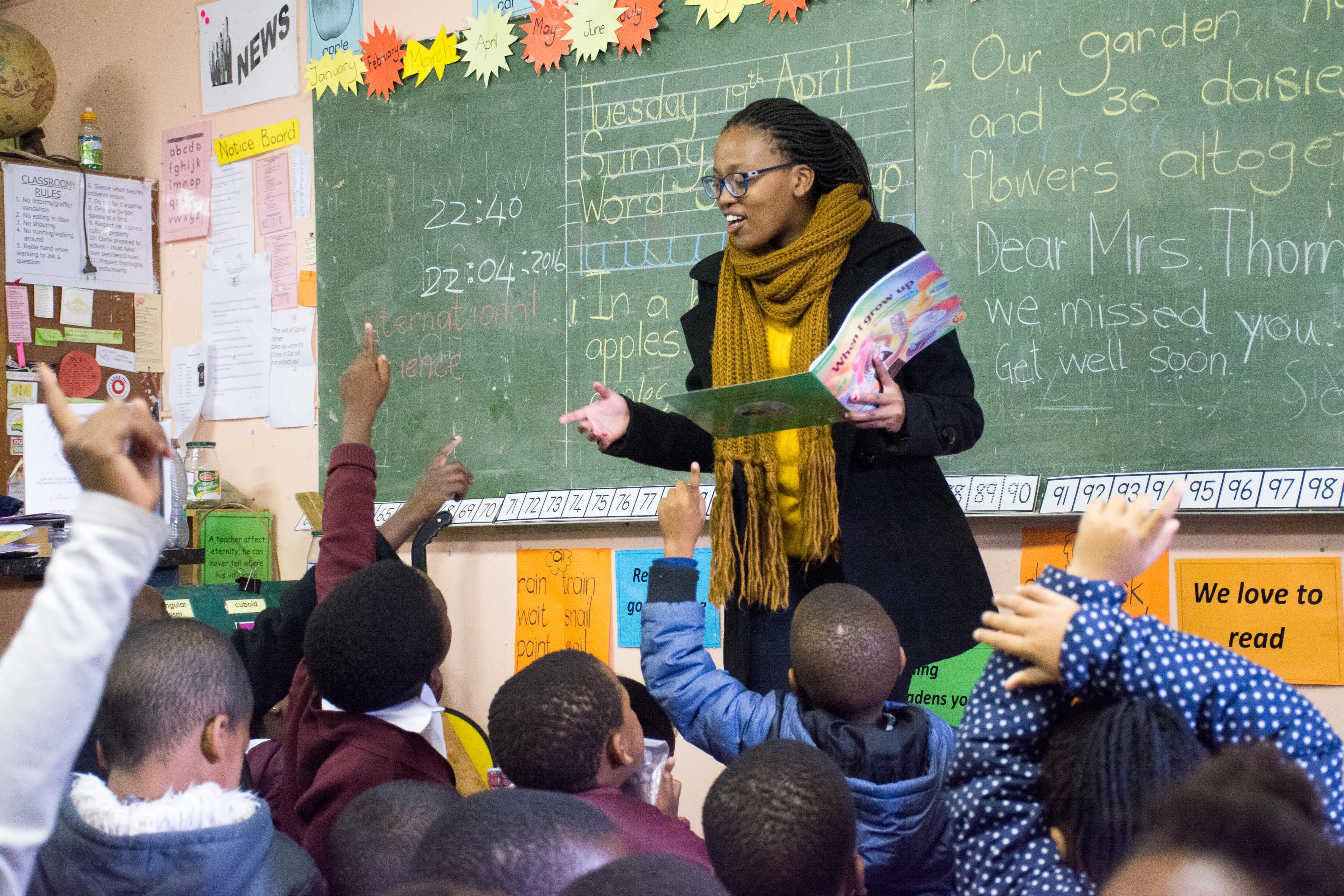A teacher stands infront of a class who have their hands in the air