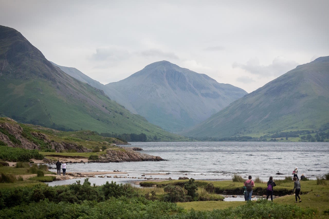 Lake District a shot of water surrounded by three hills. people are walking by the lake.